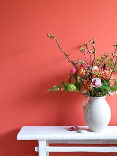 a vase with flowers sitting on a table against a pink wall in front of a white bench