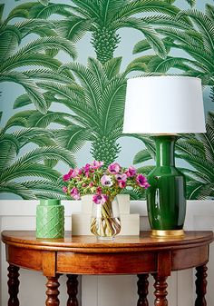 a green vase with pink flowers on a table next to a lamp and wallpaper