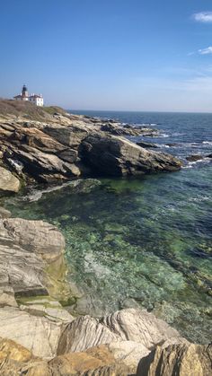 the water is crystal green and clear by the rocky shore with a lighthouse in the distance