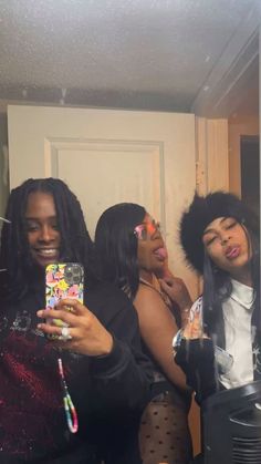 three women taking selfies in the mirror with their cell phones and one is wearing an afro wig