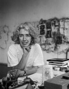 a woman sitting at a desk in front of a wall with pictures on it and various other items around her