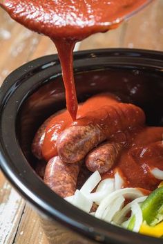 a person pouring sauce on hot dogs in a crock pot with onions, peppers and jalapenos