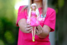 a woman holding a barbie doll in her hands and wearing a pink t - shirt