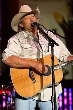 a man wearing a cowboy hat and holding a guitar in front of a microphone on stage