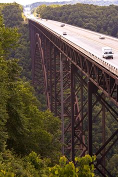 two cars are driving over a bridge in the middle of trees and mountains, with one car on it's side