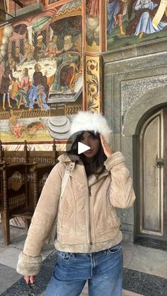 a woman standing in front of a wall with paintings on it and wearing a fur hat