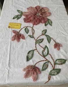 an embroidered table cloth with pink flowers and green leaves on white fabric, next to a yellow tag