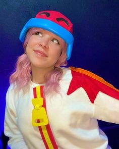 a woman with pink hair wearing a white shirt and red and yellow hat, standing in front of a blue background