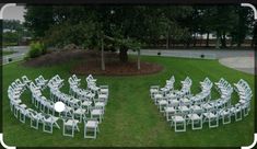 an arrangement of white chairs arranged in the shape of a circle on green grass near a tree