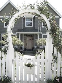 a white picket fence in front of a house with a wreath on the gate and flowers around it