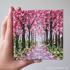 a hand holding up a small card with a painting of trees and flowers on it