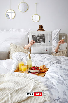 a woman reading a newspaper while laying in bed with fruit and juice on the tray