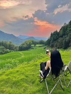 a woman sitting in a lawn chair taking a photo with her cell phone at sunset