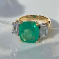 an emerald and diamond three stone ring on a white surface with other jewelry items in the background