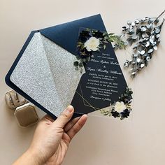 a hand holding an open black and silver wedding card with white flowers on the inside