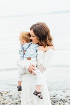 a woman holding a baby in her arms while standing on the beach with water behind her