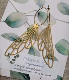 a pair of gold - toned metal earrings with leaves on them are displayed in front of a thank card