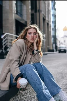 a woman is sitting on the curb with her hand under her chin and holding a coffee cup