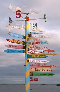 a pole with many signs on it in front of the ocean and cloudy sky above