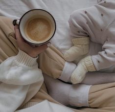 a person holding a cup of coffee while laying on top of a white bed covered in blankets