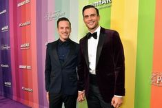 two men standing next to each other in front of a rainbow colored wall wearing tuxedos