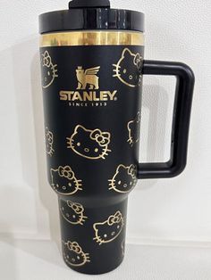 a black and gold travel mug with hello kitty designs on it's side, next to a white wall