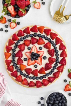 a pie topped with strawberries and blueberries on top of a white table next to utensils