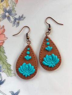 two pairs of brown and blue leather earrings on a white surface with flowers in the background