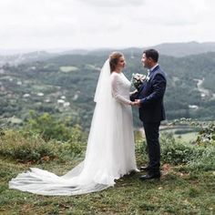 a bride and groom standing on top of a hill looking at each other while holding hands