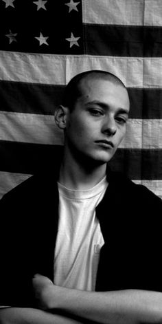 black and white photograph of a man in front of an american flag with his arms crossed