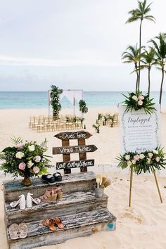 a wooden sign sitting on top of a sandy beach next to flowers and palm trees