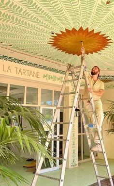 a woman is painting the ceiling in a room with palm trees and potted plants