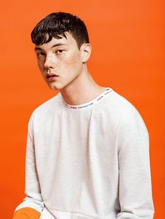 a young man with freckled hair wearing a white shirt and orange gloves, standing in front of an orange background