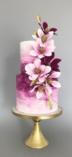 a three tiered cake with pink and purple flowers on the top, sitting on a gold plate