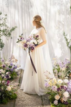 a woman in a white dress holding a bouquet and standing next to some purple flowers