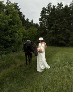 a woman in a white dress and hat walking with a black horse on a path