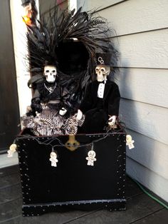 two skeletons are sitting in a trunk on the porch with halloween decorations and fake feathers