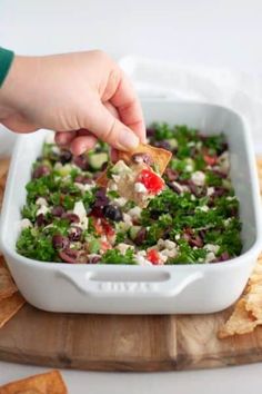 a person dipping tortilla chips into a white casserole dish filled with black olives, cilantro, and red pepper