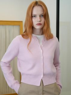 a woman with red hair wearing a pink cardigan and khaki pants standing in front of a mirror