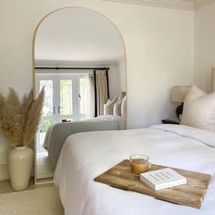 a bed with white sheets and pillows in a bedroom next to a mirror that has a book on it