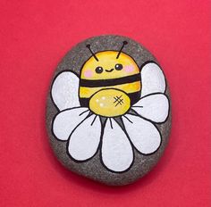 a painted rock with a bee on it sitting on a red surface next to a yellow and white flower