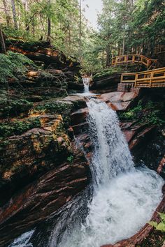 a waterfall in the middle of a forest with a wooden bridge above it and trees around