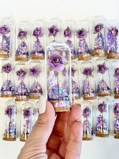 a hand holding a tiny glass tube with purple flowers inside it and lots of other small bottles in the background