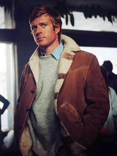 a man wearing a jacket and sweater standing in front of a window