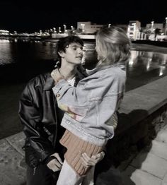 a man and woman standing next to each other on a sidewalk near the water at night