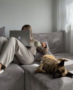 a woman sitting on a couch holding a laptop computer and drinking coffee with her dog