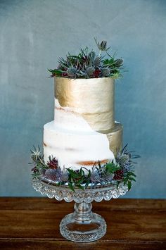 a three tiered cake with pine cones and greenery on top sits on a glass pedestal