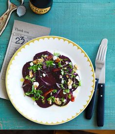 a white plate topped with beet salad next to a fork and knife on top of a blue table