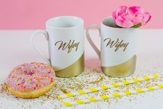 two coffee mugs sitting next to a doughnut on a pink surface with sprinkles