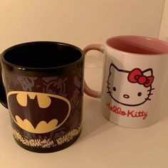 two coffee mugs with hello kitty and batman designs on them, one has a red bow